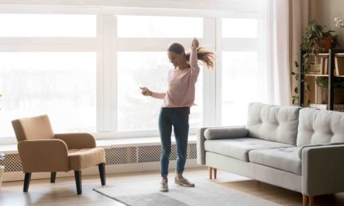 Happy carefree young woman dancing alone having fun at home listening to good music, energetic girl moving jumping in modern living room interior with large window enjoy freedom and active lifestyle